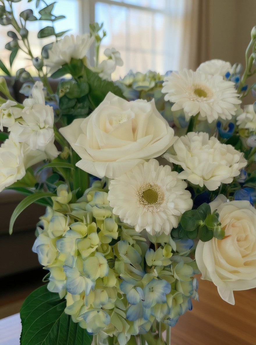 Lakewood Florist Crisp Sky arrangement: white roses, gerbera daisies, blue delphiniums, and hydrangeas with greenery in a clear vase.